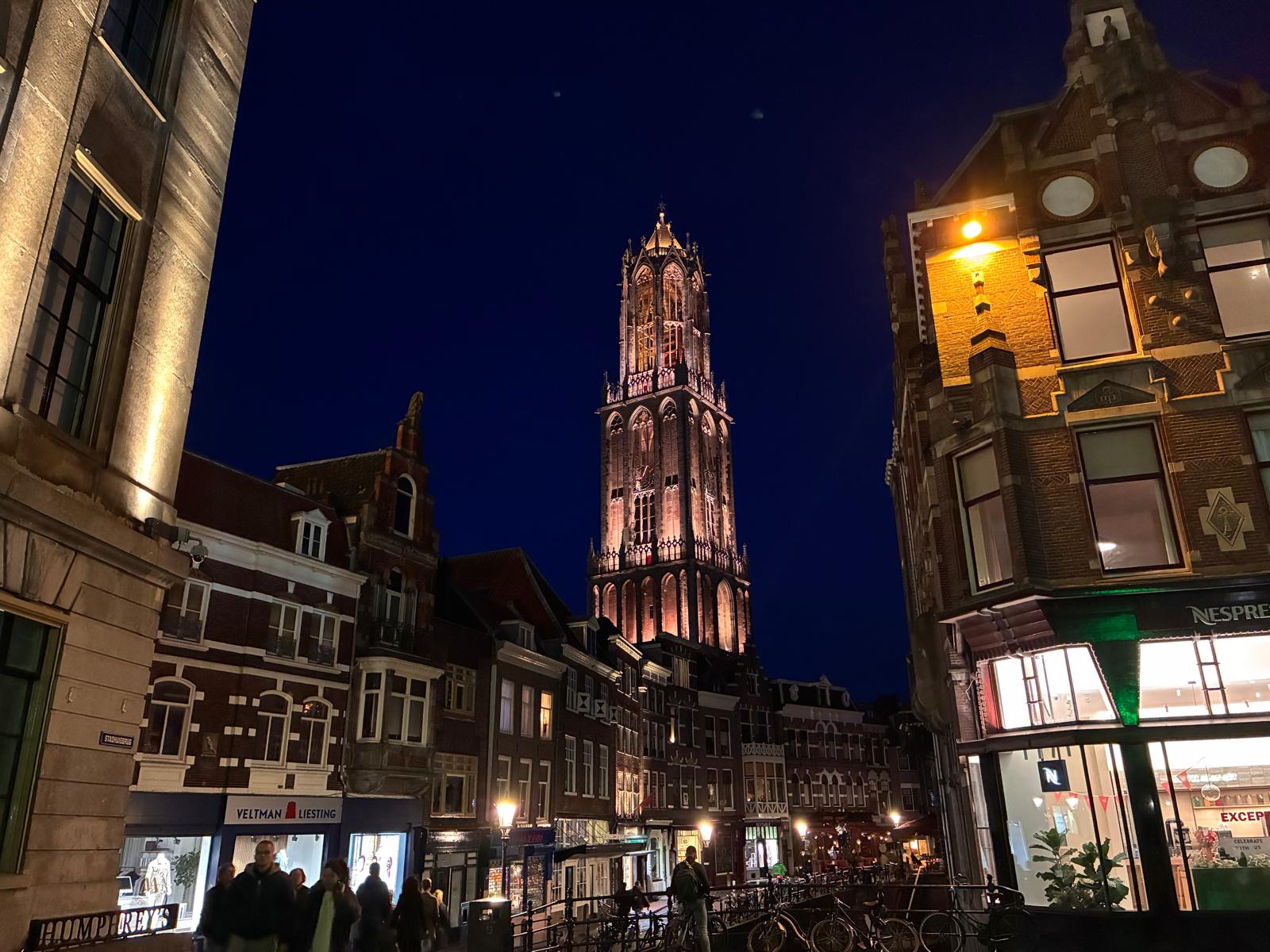 The illuminated Dom Tower of Utrecht at night, viewed from the Stadhuisbrug with historic canal houses and shops along the Oudegracht